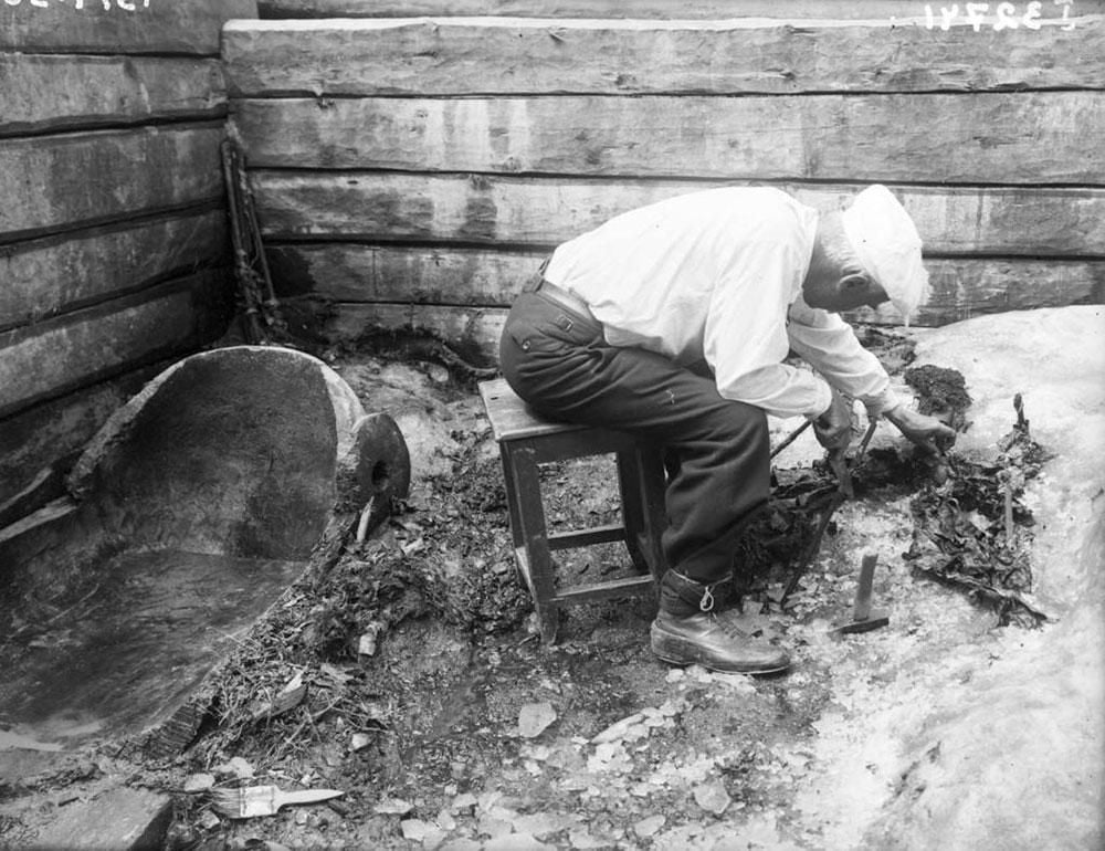 An archaeologist, sitting on a stool, carefully excavates a tomb chamber which is buried under ice